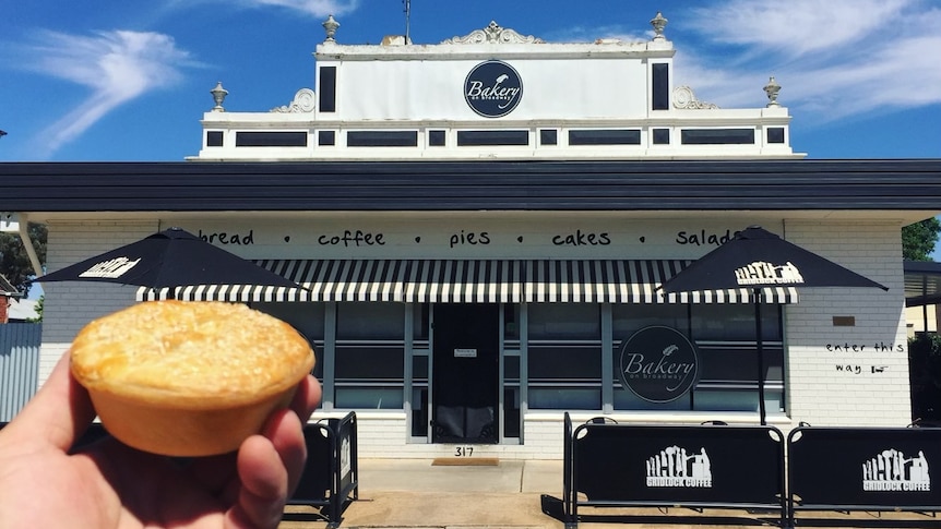 Party pie held in a hand out the front of a white bakery building with black outdoor dining facilities