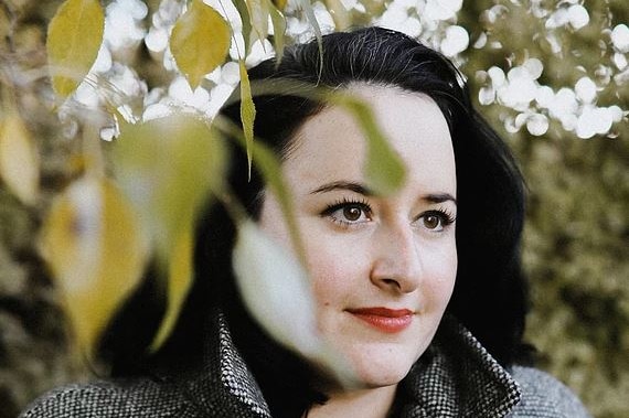 A young woman with dark hair stands behind some tree leaves and looks off to the side of the frame.