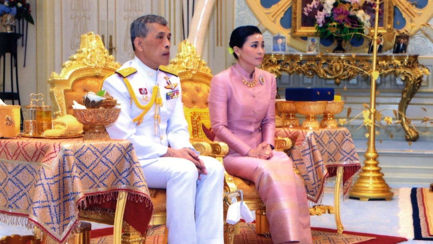 Thailand's king and queen sit side by side in a palace room, decorated in yellow and gold.