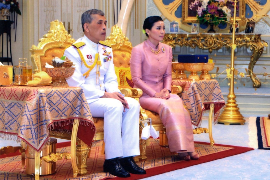 Thailand's king and queen sit side by side in a palace room, decorated in yellow and gold.