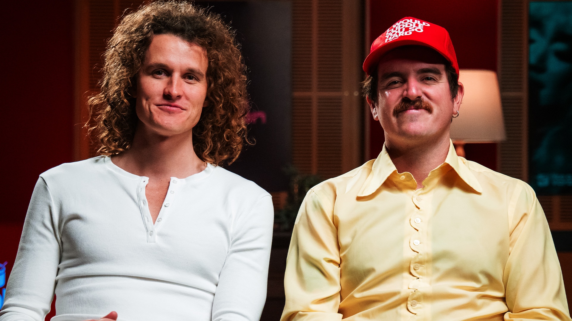 A man with curly hair in a white longsleeve top and a man in a red trucker hat and a 1970s yellow shirt smile at the camera
