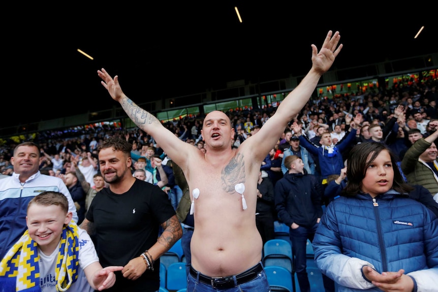 A rotund, bald, tattooed, middle aged man with love heart-shaped nipple tassels fixed to his chest holds his arms up.