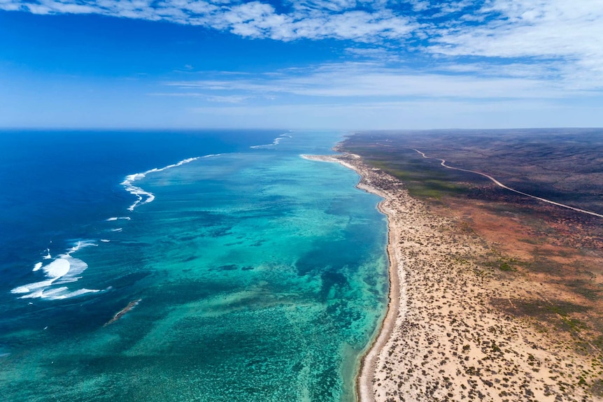 Aerial of bright blue sea and sandy and red dirt shoreline.