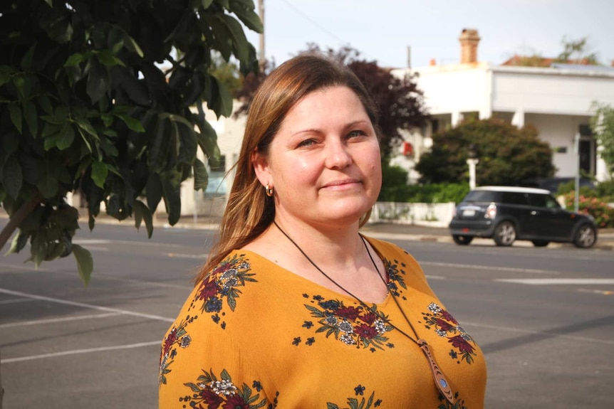 A woman stands on a suburban street, she is wearing a yellow floral top.