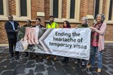 A group of people standing outside holding a banner that says "Ending the heartache of temporary visas".