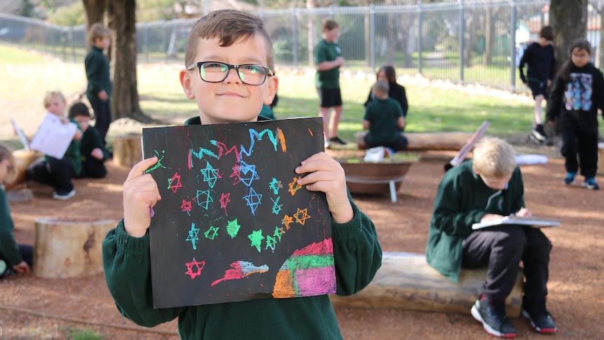 A young boy in school uniform holds up a drawing he completed in the yarn circle.