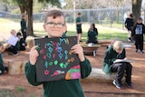 A young boy in school uniform holds up a drawing he completed in the yarn circle.