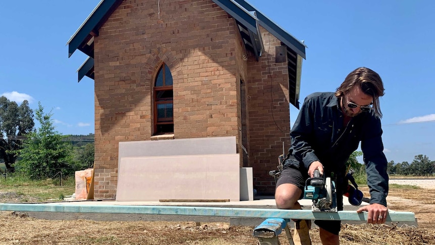 A young man sawing a piece of timber in front of a building in a rural town.