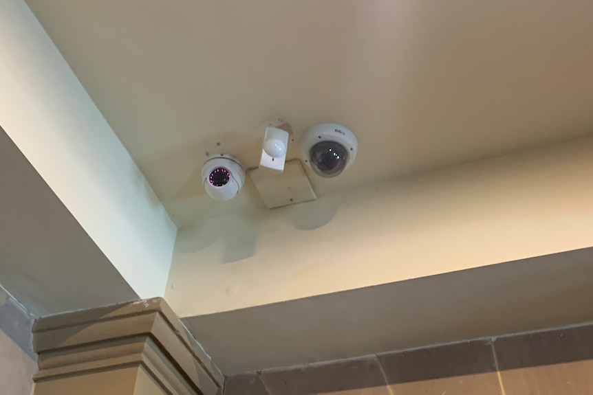 Two security cameras are seen in the upper corner of a room near the ceiling.