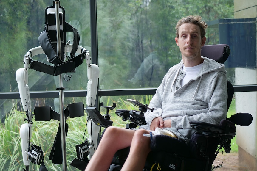 A young man wears a grey jumper while sitting in his wheelchair, alongside an exoskeleton.