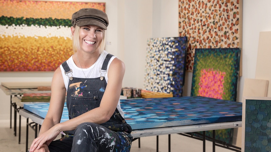 A woman dressed in overalls sitting on a stool in an art studio.