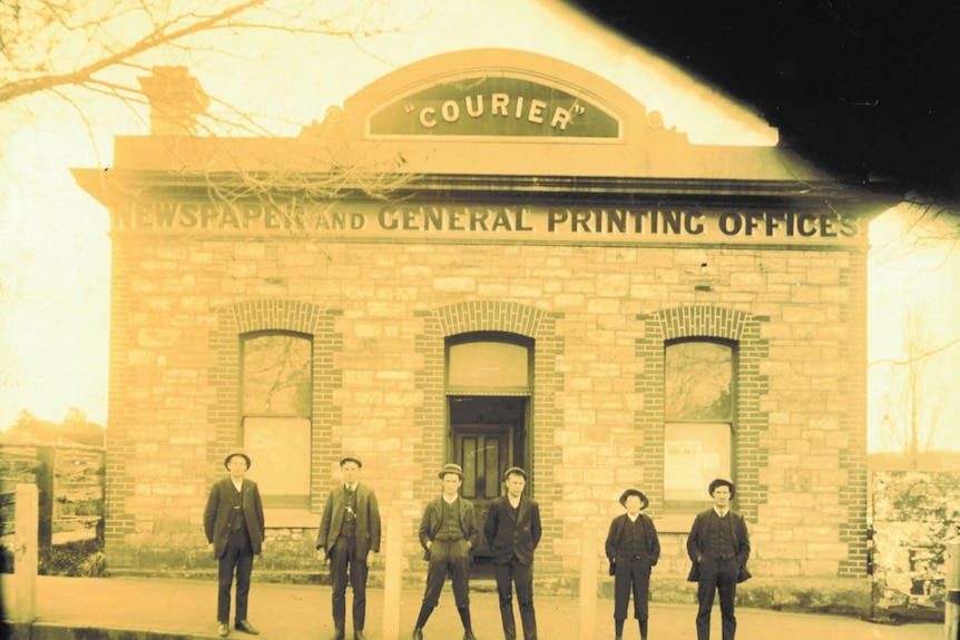 An old, yellowed photo of a group of men standing outside a brick building with a sign that reads "COURIER".