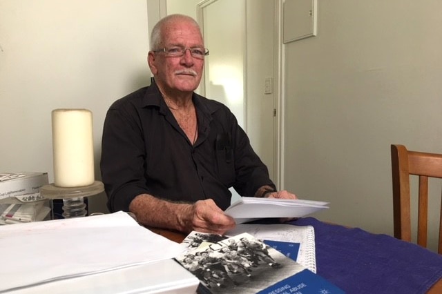 An elderly man wearing glasses  sits a table with a number of books, folders and documents.