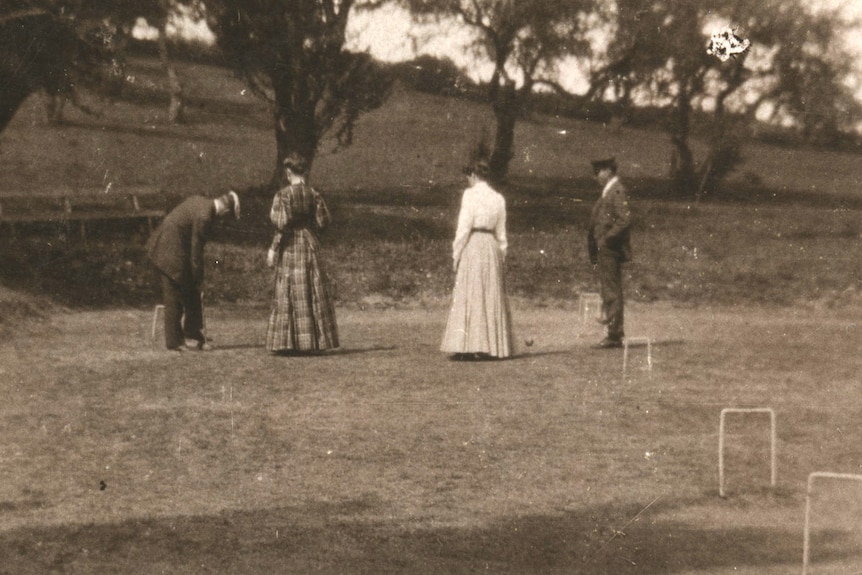 Two men and two women dressed in the clothes of early 20th century play croquet in a park.