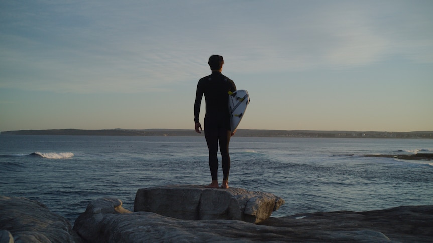 A man in a wetsuit and carrying a surfboard stands on a rock looking out to the sea at sunrise