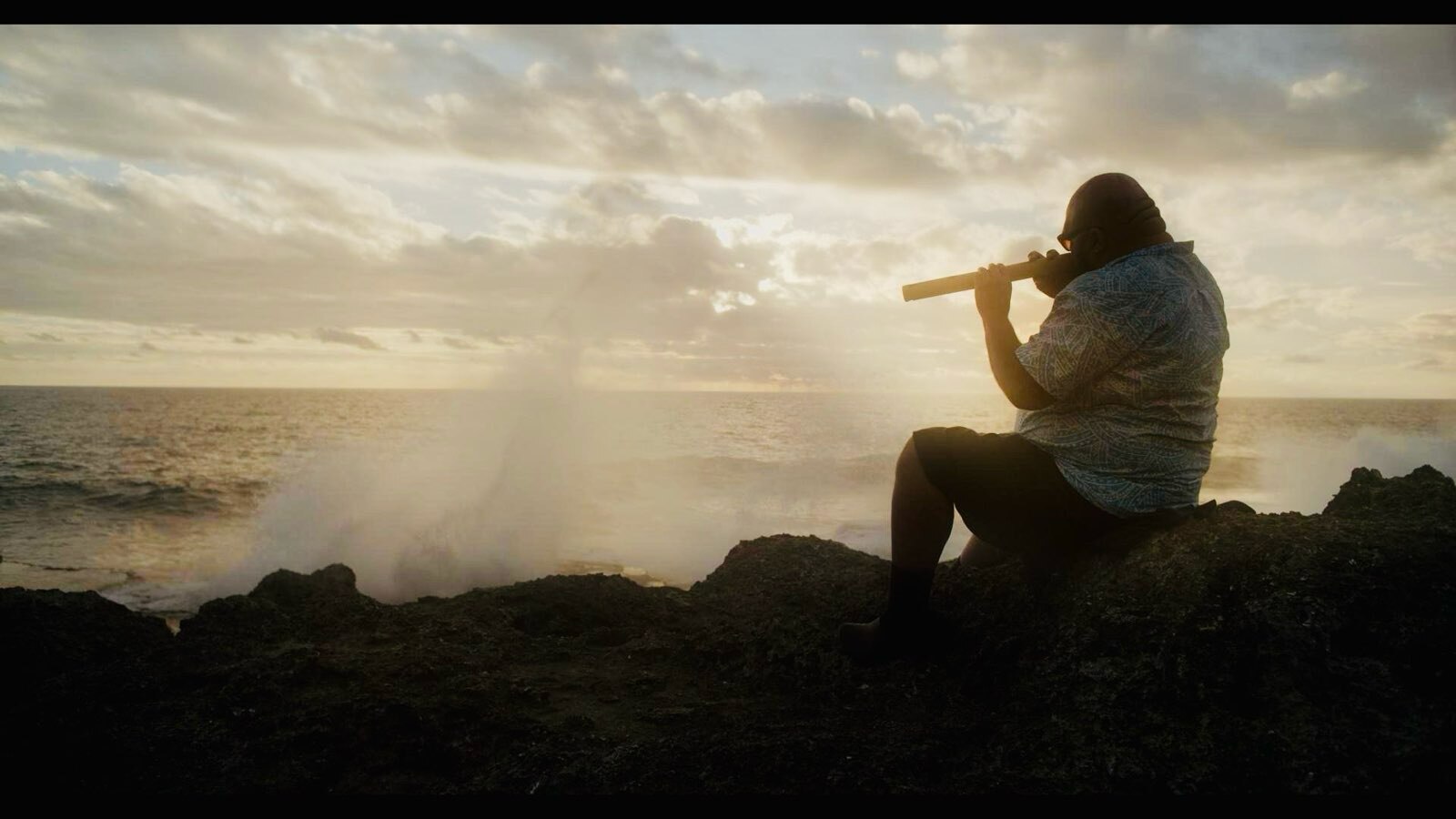 Man plays instrument by ocean as sun sets.