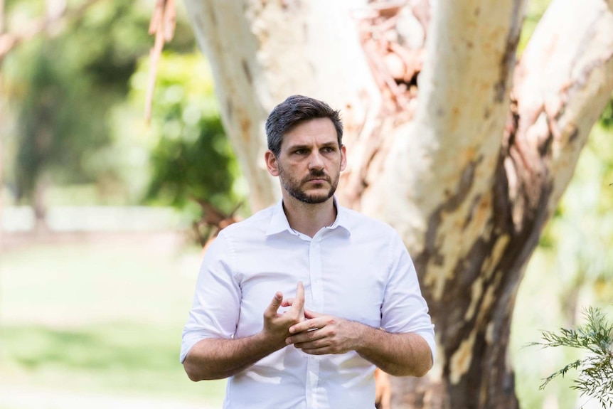 A dark-haired man with neat facial hair, wearing a business shirt, stands outside and speaks to the media.