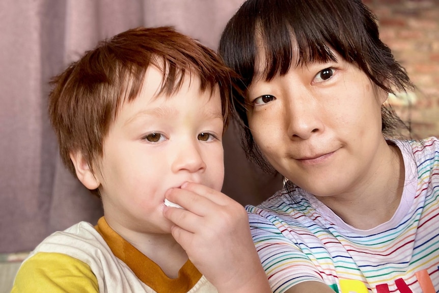 Woman smiles next to toddler as he puts food in his mouth. 