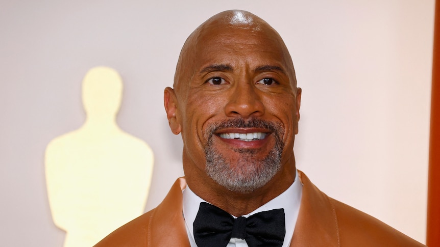 An image of Dwayne 'The Rock' Johnson wearing a brown jacket with a white shirt and a black bowtie.