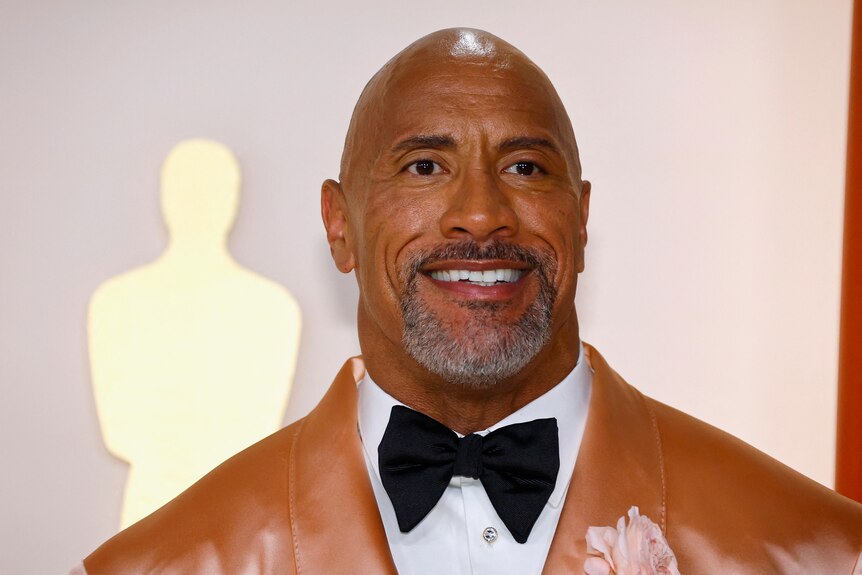 An image of Dwayne 'The Rock' Johnson wearing a brown jacket with a white shirt and a black bowtie.