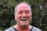 Barnaby Joyce, sweaty and red in the face, crinkles his eye shut while laughing. Microphones are visible in front of him.