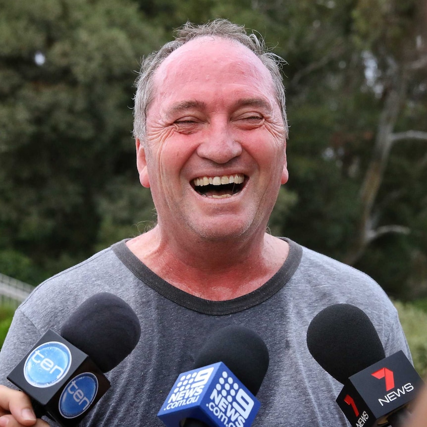 Barnaby Joyce, sweaty and red in the face, crinkles his eye shut while laughing. Microphones are visible in front of him.