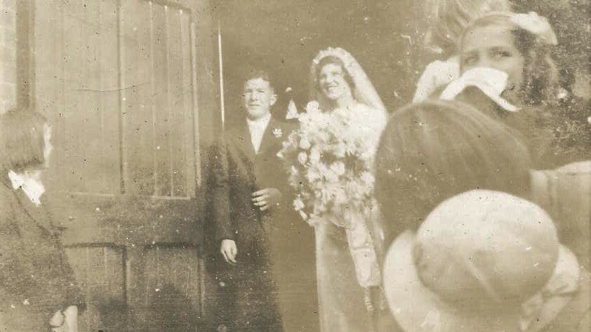 A vintage photograph in faded sepia shows a bride and groom walking through a crowd at the church door. 