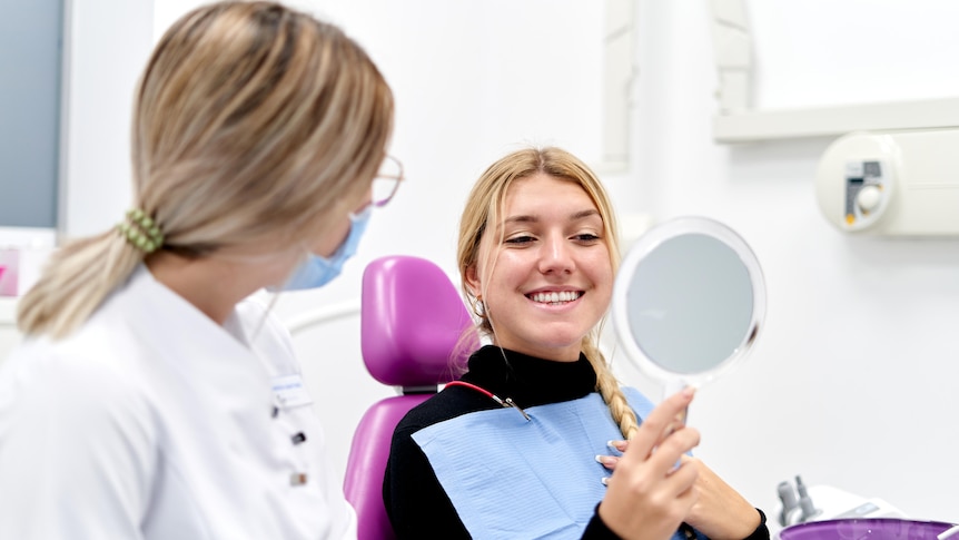 A young woman smiles into a mirror while a female hygienist looks on