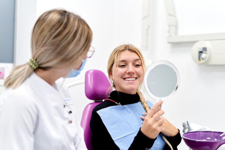 A young woman smiles into a mirror while a female hygienist looks on