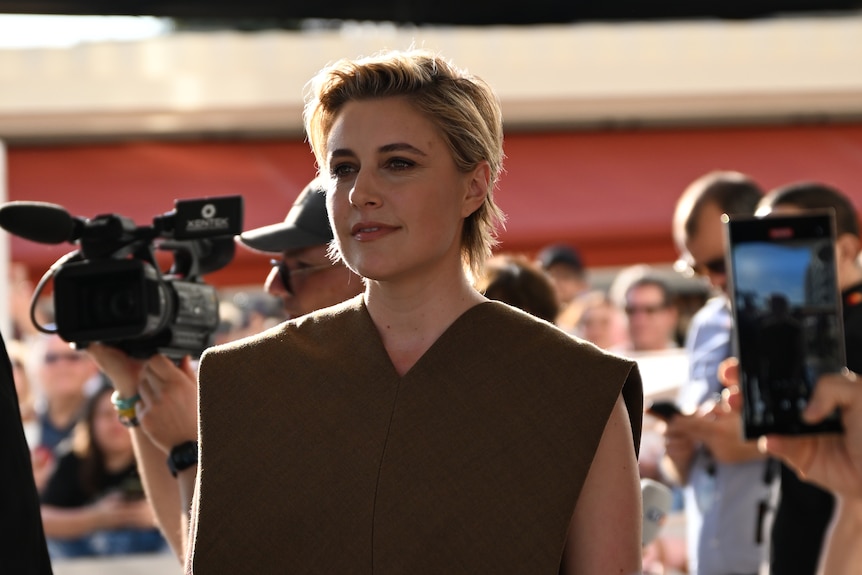 A woman in a brown vneck dress stands in front of a sea of people, with cameras of all size - phone, TV - around.