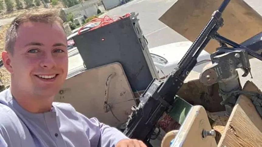 A young white male with short hair smiling in selfie with mounted gun pictured in the background.