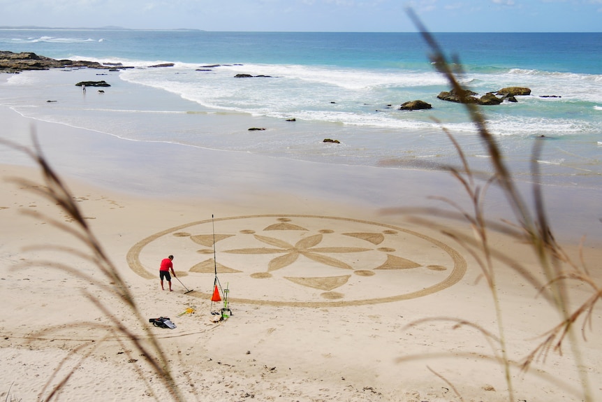 A man with a rake creates a geometric design on the sand on a beach.