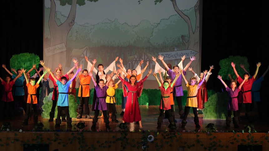 A group of young actors in Robin Hood costumes raise their hands in the air while performing on stage.