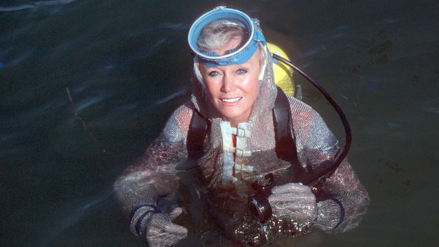 A woman with blonde hair floating in the ocean wearing a mesh scuba suit with a diving mask on top of her head.