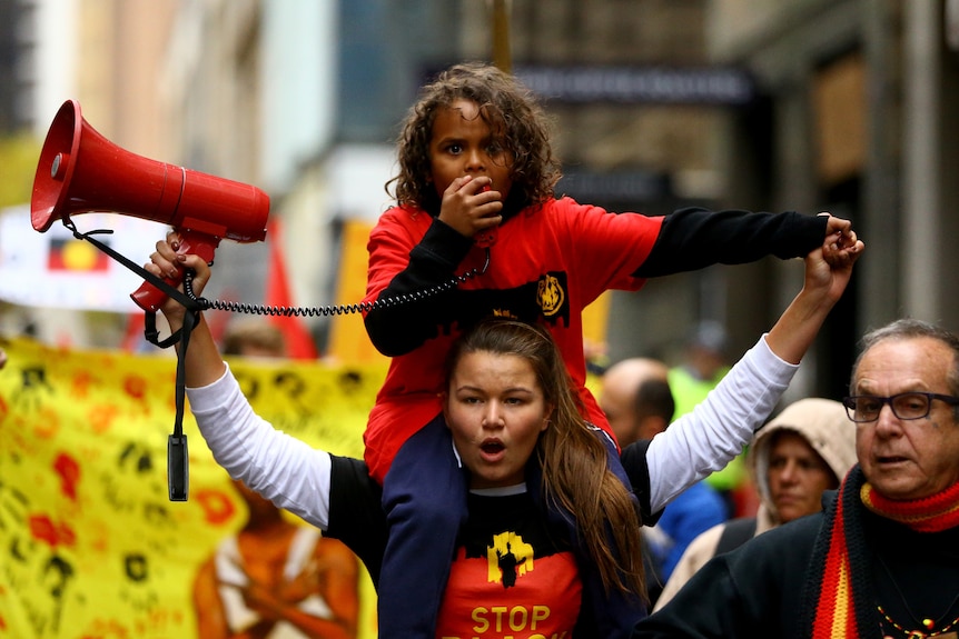 Vanessa Turnbull-Roberts at a rally, holding a megaphone in one hand and carrying a child on her shoulders.