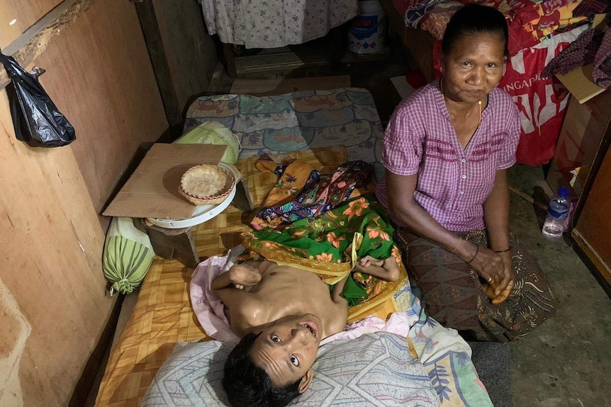 A mother sits by her son's bed in their home, both looking at camera.
