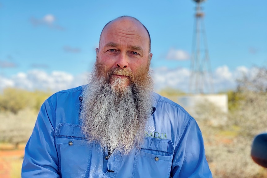 A man in blue with a beard looks at the camera, a windmill in the background.