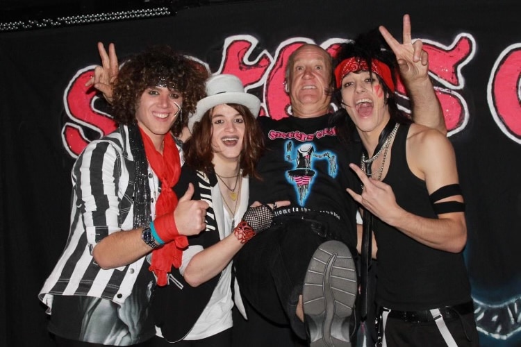 An older man wears a Sisters Doll t-shirt, his arms are around three band members in glam rock attire, all smiling.