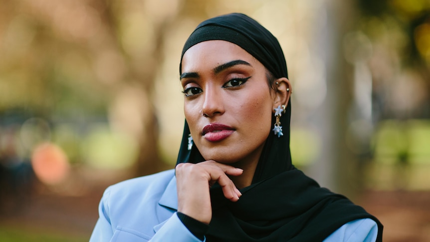 Young Muslim woman Maab wears sky blue jacket and black hijab, with her hand resting below her chin.