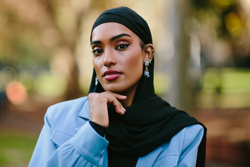 Young Muslim woman Maab wears sky blue jacket and black hijab, with her hand resting below her chin.