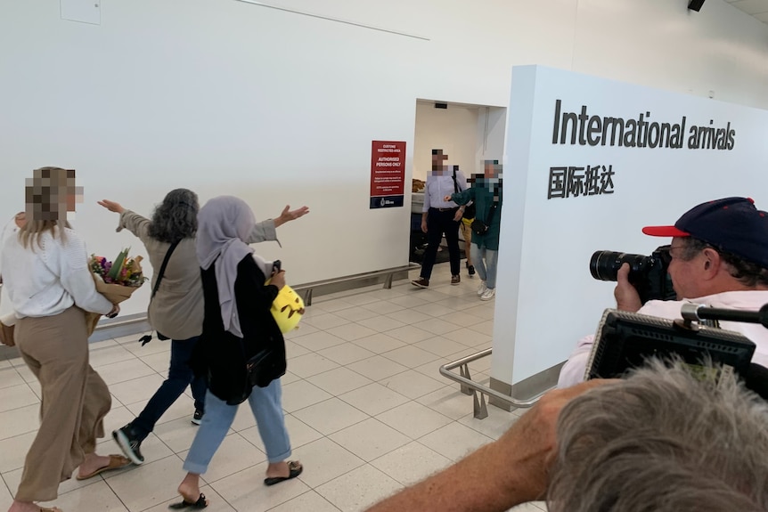 A woman and man walking inside an aiport being greeted by three women with flowers. Faces are pixelated. 