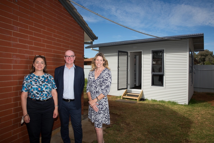 Two women and a man stand in front of a granny flat in a backyard, behind a house