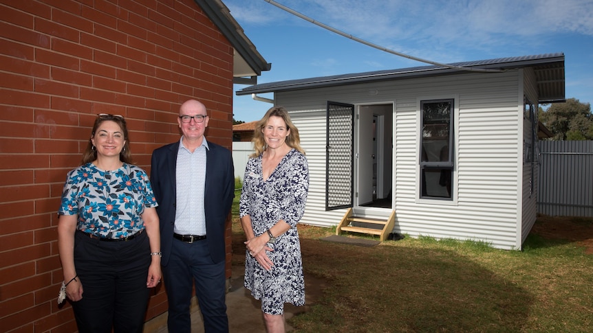 Two women and a man stand in front of a granny flat in a backyard, behind a house