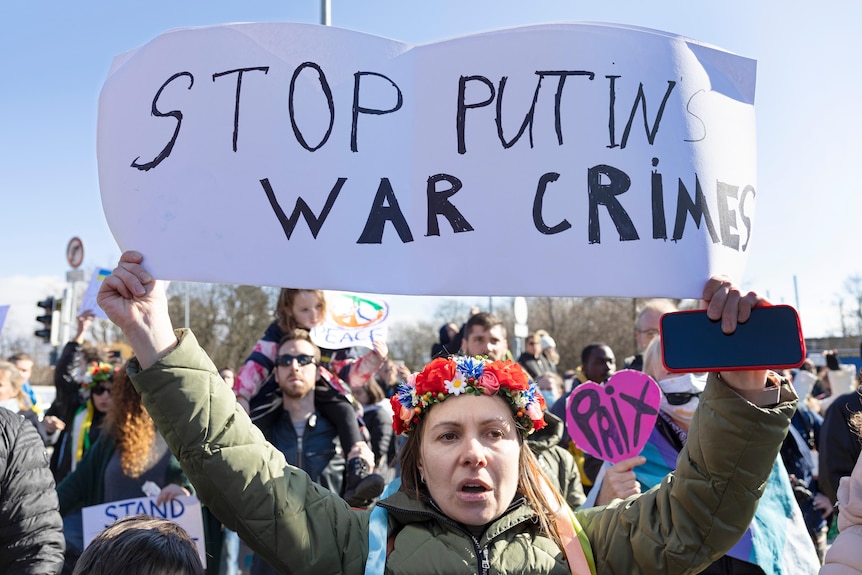 한 시위자가 다음과 같은 표지판을 들고 있습니다. "푸틴의 전쟁 범죄를 중지" 많은 군중 앞에 섰을 때.