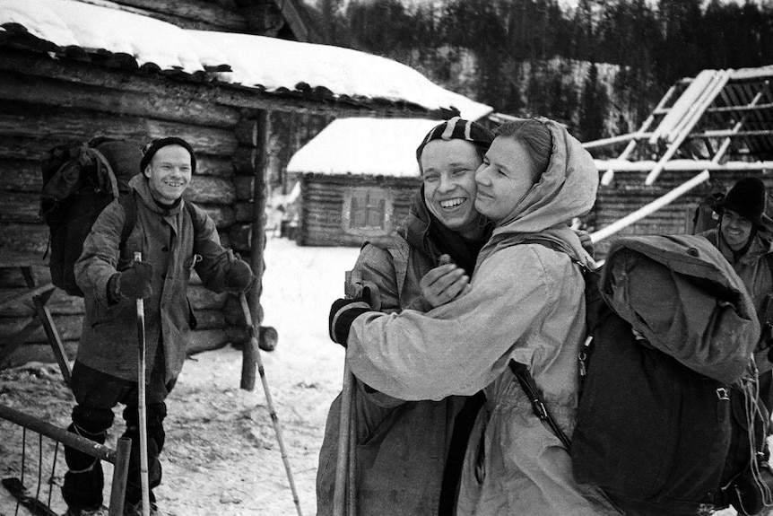 A black and white photo of a man and a woman embracing while another man with ski poles looks on and laughs 