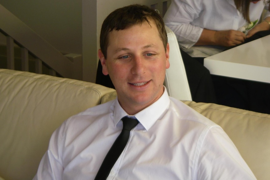 A man in a shirt and tie sits on a couch. A person sits in a dining chair in the background