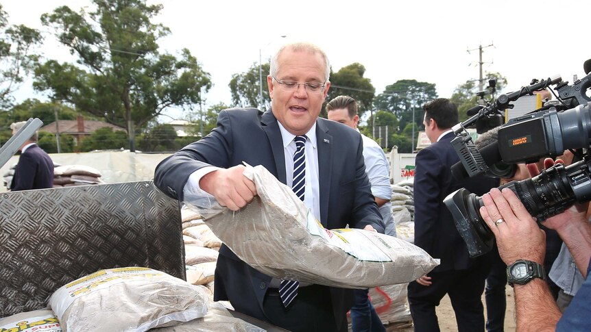 Scott Morrison, wearing a suit, looks at a bag of sand as he lifts it into the trailer of a truck