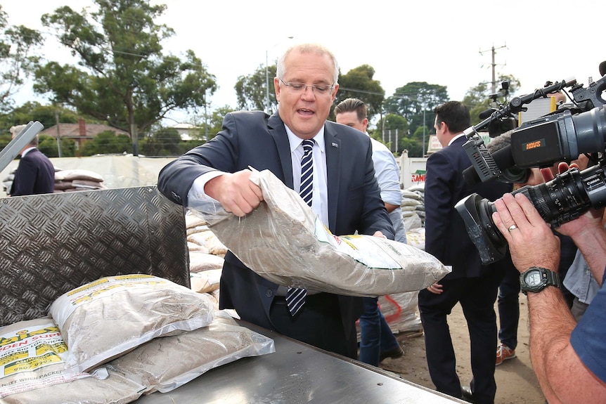 Scott Morrison, wearing a suit, looks at a bag of sand as he lifts it into the trailer of a truck