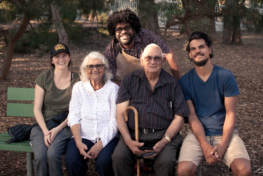 Two elderly people on a park bench are flanked by one man and a woman, while a man with a beard stands behind smiling.