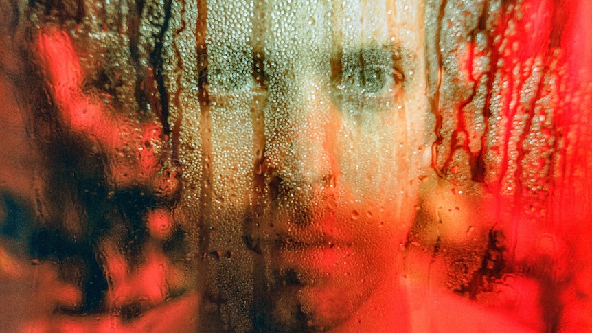 Jay Watson's face is distorted behind a window with water running down it